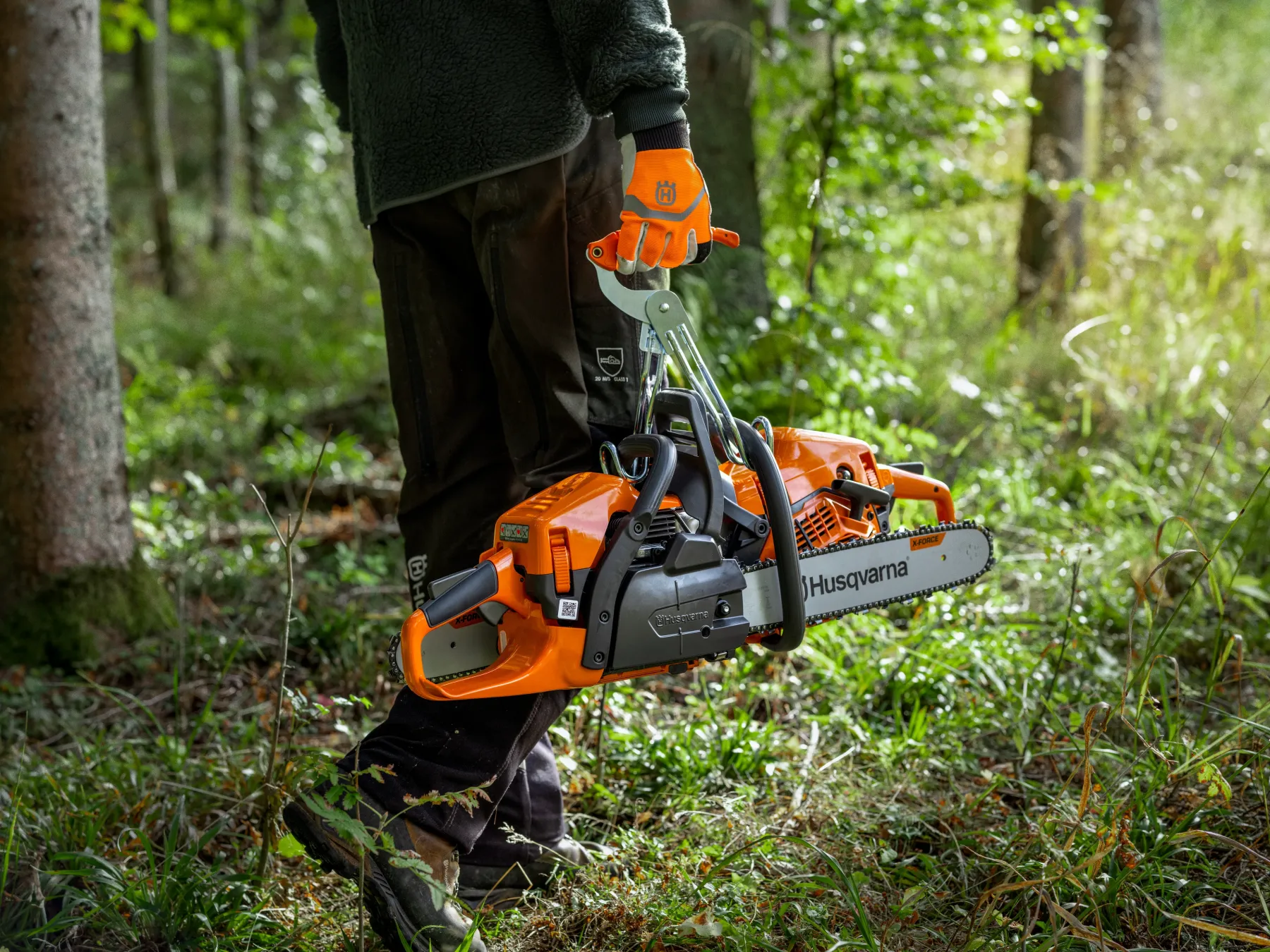 En person iförd handskar och friluftskläder bär två orange Husqvarna-motorsågar i en grön skog och går genom gräs och träd.