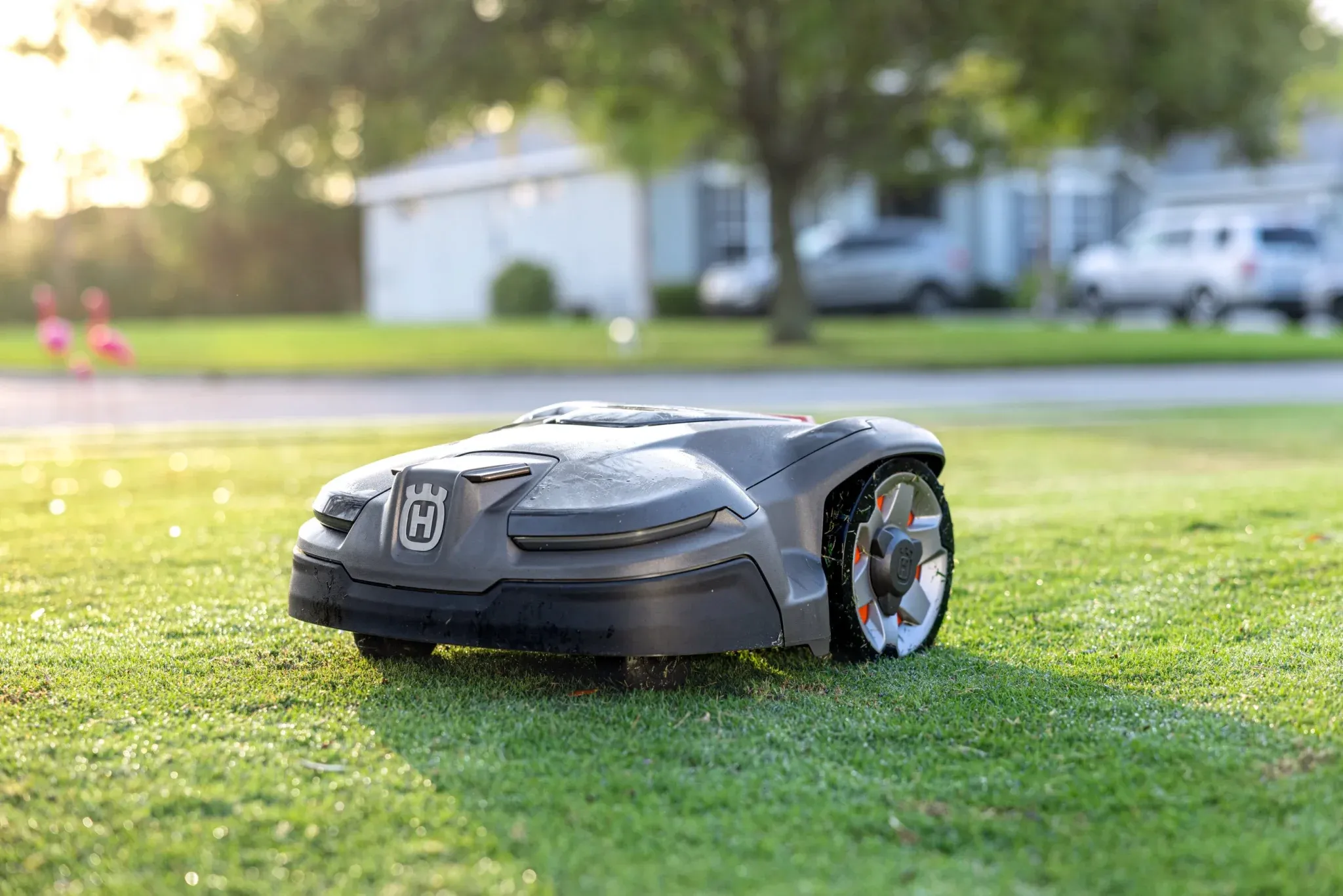 En robotgräsklippare med en elegant svart design rör sig över en gräsmatta i ett förortsområde, med träd, en väg och parkerade bilar i bakgrunden.