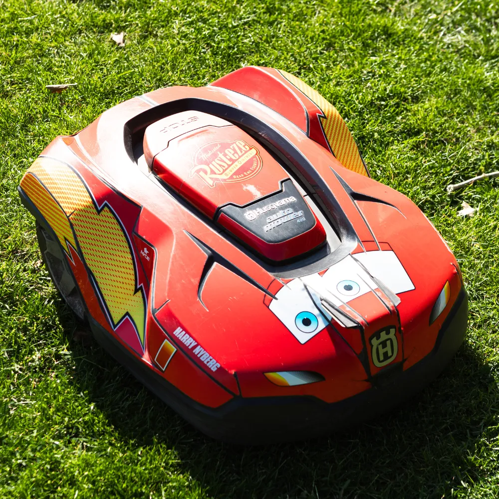 En personlig robotgräsklippare, utsmyckad som Blixten McQueen från Bilar med tecknade ögon och racingdekaler, sitter på grönt gräs i starkt solljus.