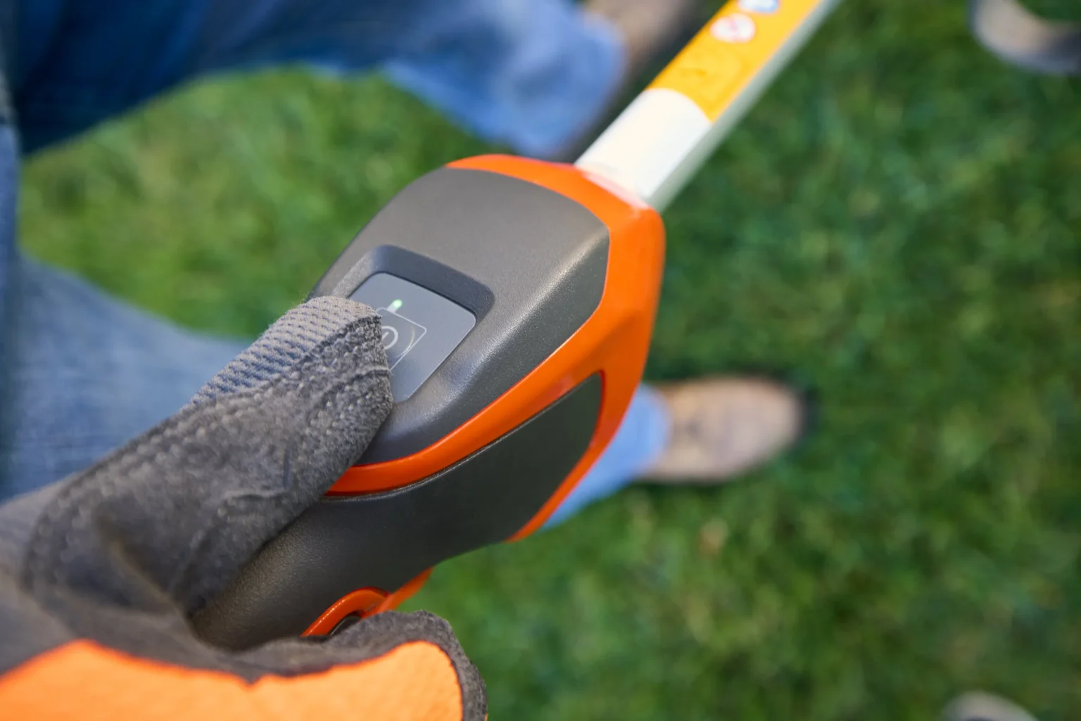 Grästrimmer med batteri En handskbeklädd hand trycker på strömbrytaren på handtaget till ett orange och svart elverktyg, med grönt gräs och en persons fot synlig i den suddiga bakgrunden.