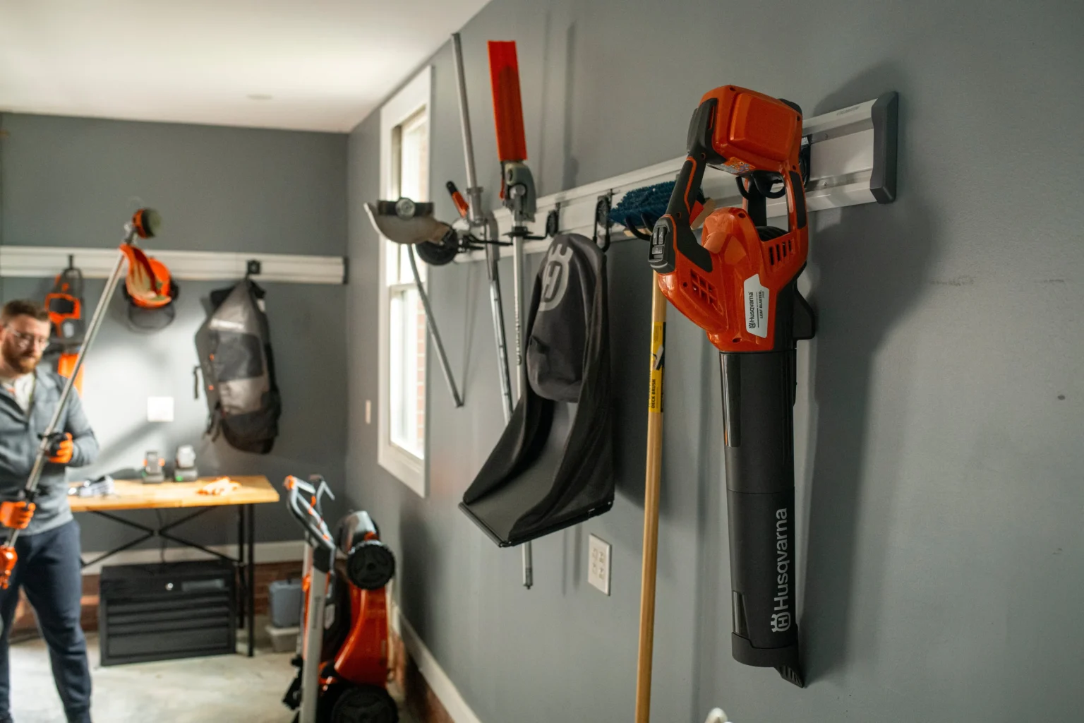 Service - robotgräsklippare Ett garage med orange och svarta Husqvarna trädgårdsredskap och utrustning, inklusive en robotgräsklippare för idrottsföreningar, organiserade på väggmonterade ställningar. En person håller i en trimmer i bakgrunden nära en arbetsbänk med trädgårdsredskap.