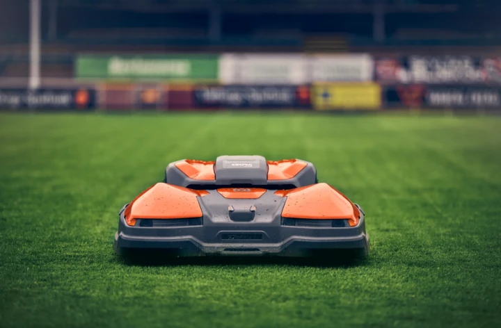 En orange och grå robotgräsklippare sitter på nyklippt gräs på en stadion, med suddiga reklamskyltar synliga i bakgrunden.