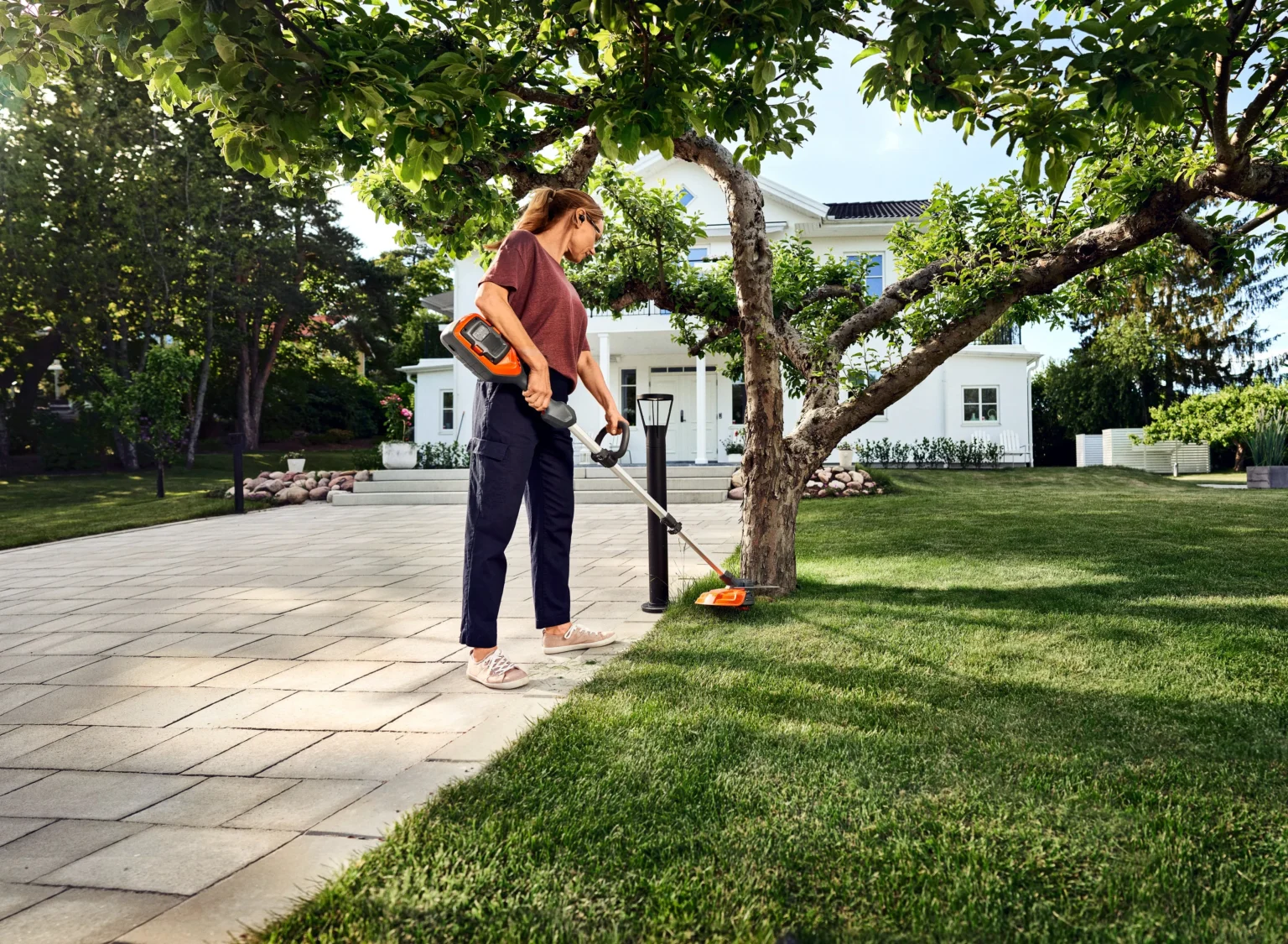 Grästrimmer - ergonomi En kvinna använder en sladdlös grästrimmer för att klippa gräsmattan nära ett träd i en solig trädgård, med ett vitt hus och en stenlagd uteplats i bakgrunden.