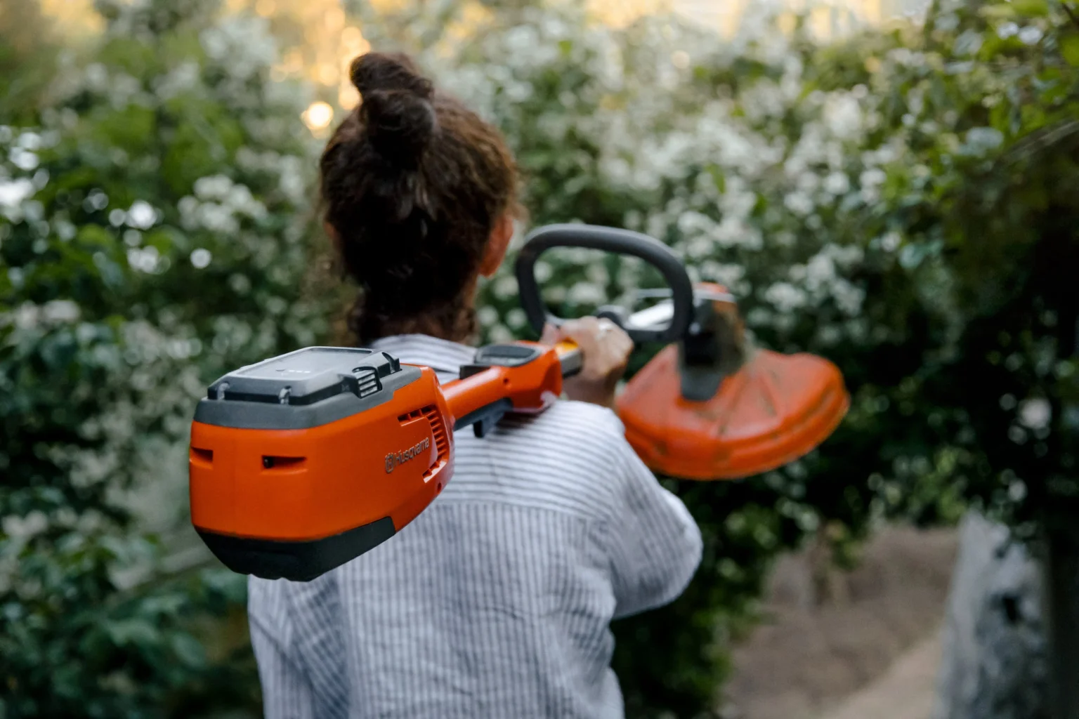Husqvarna grästrimmer En person med lockigt hår i en knut, klädd i en randig skjorta, bär en orange snortrimmer över axeln medan han går genom en trädgård med gröna buskar och vita blommor.