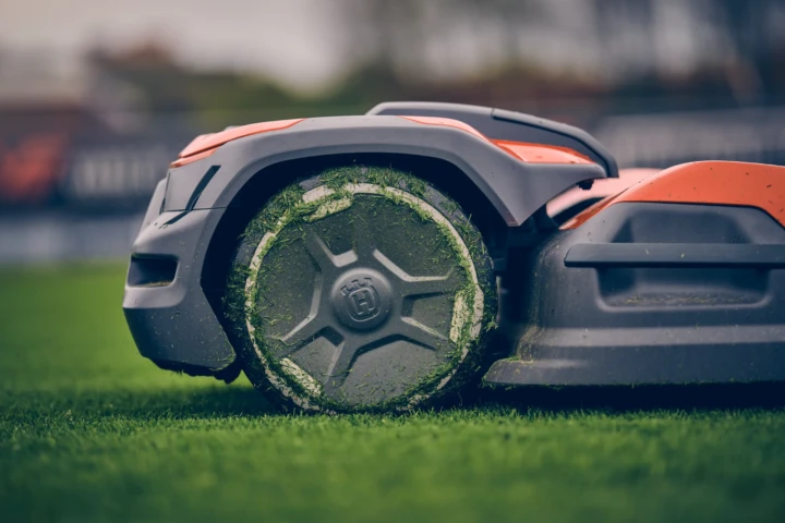 Närbild av en robotgräsklippare på grönt gräs, med gräsklipp som fastnat på hjulet. Gräsklipparen är mestadels grå med orange inslag, och bakgrunden är suddig.
