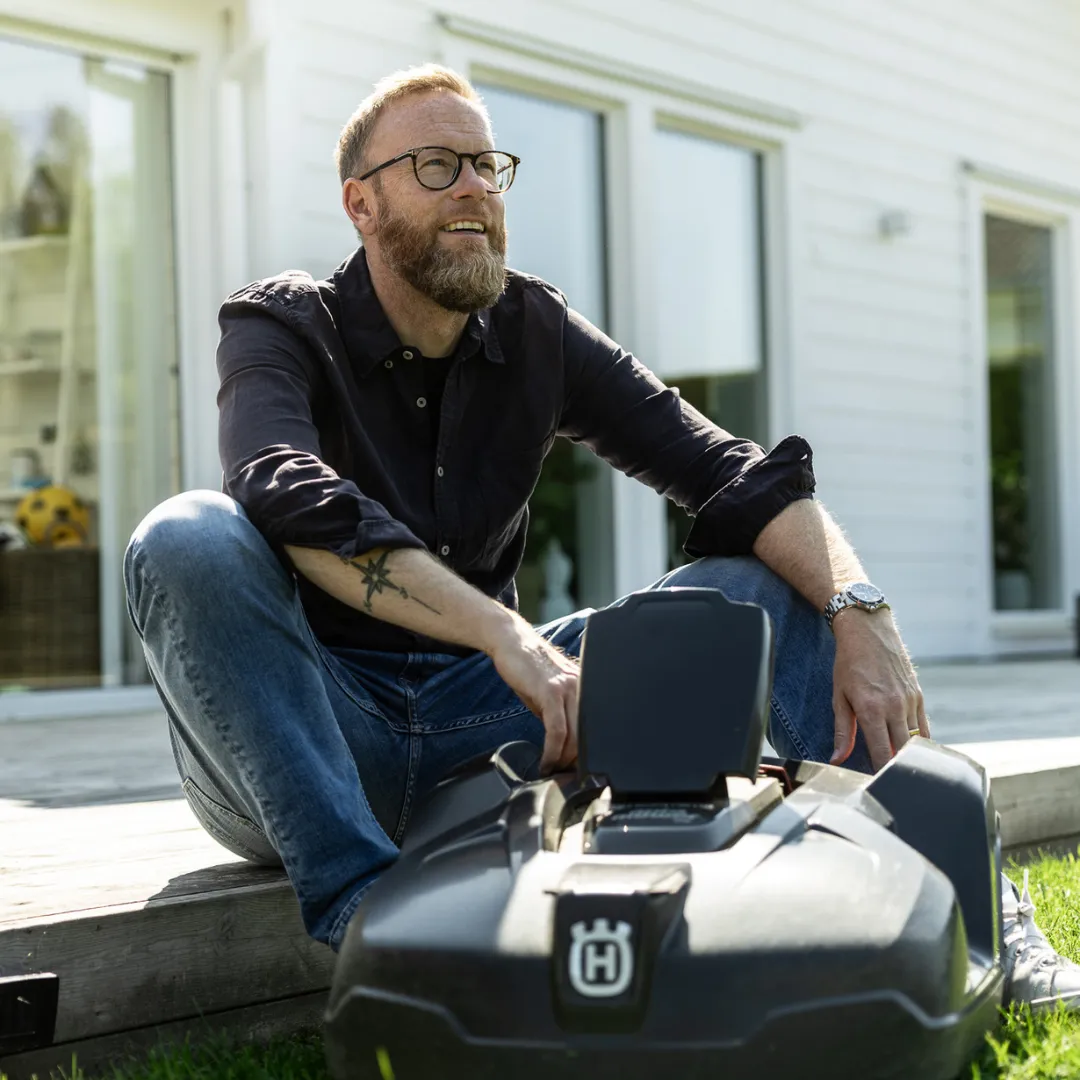 Connys Skog & Trädgård - Pierre En leende man med glasögon och skägg sitter på ett trädäck bredvid sin personliga robotgräsklippare, med ett vitt hus och en grön gräsmatta i bakgrunden.