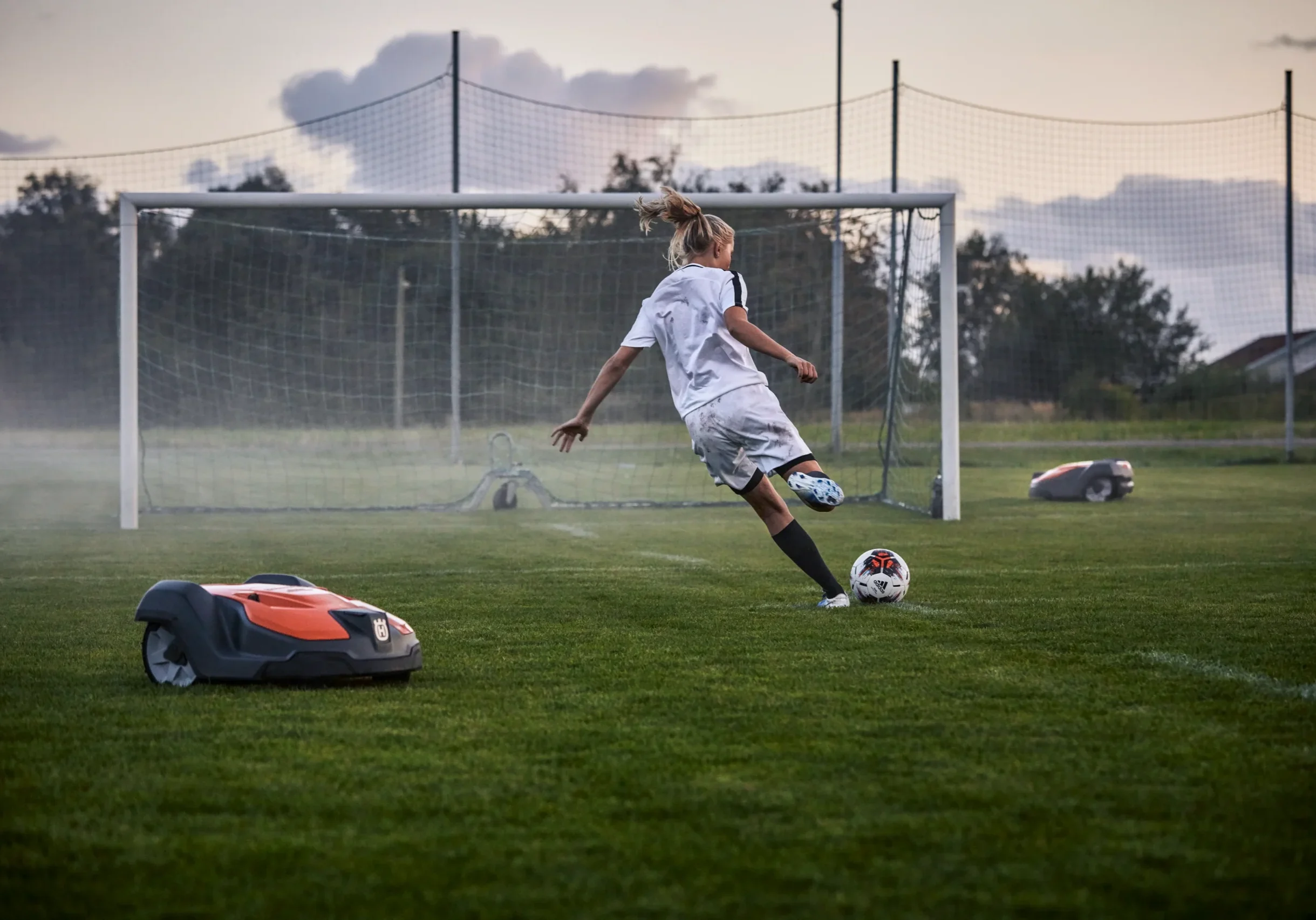 H350-0770 En ung fotbollsspelare i vit uniform sparkar en boll mot ett mål på en gräsplan, medan två robotgräsklippare lugnt sköter om gräset i närheten.
