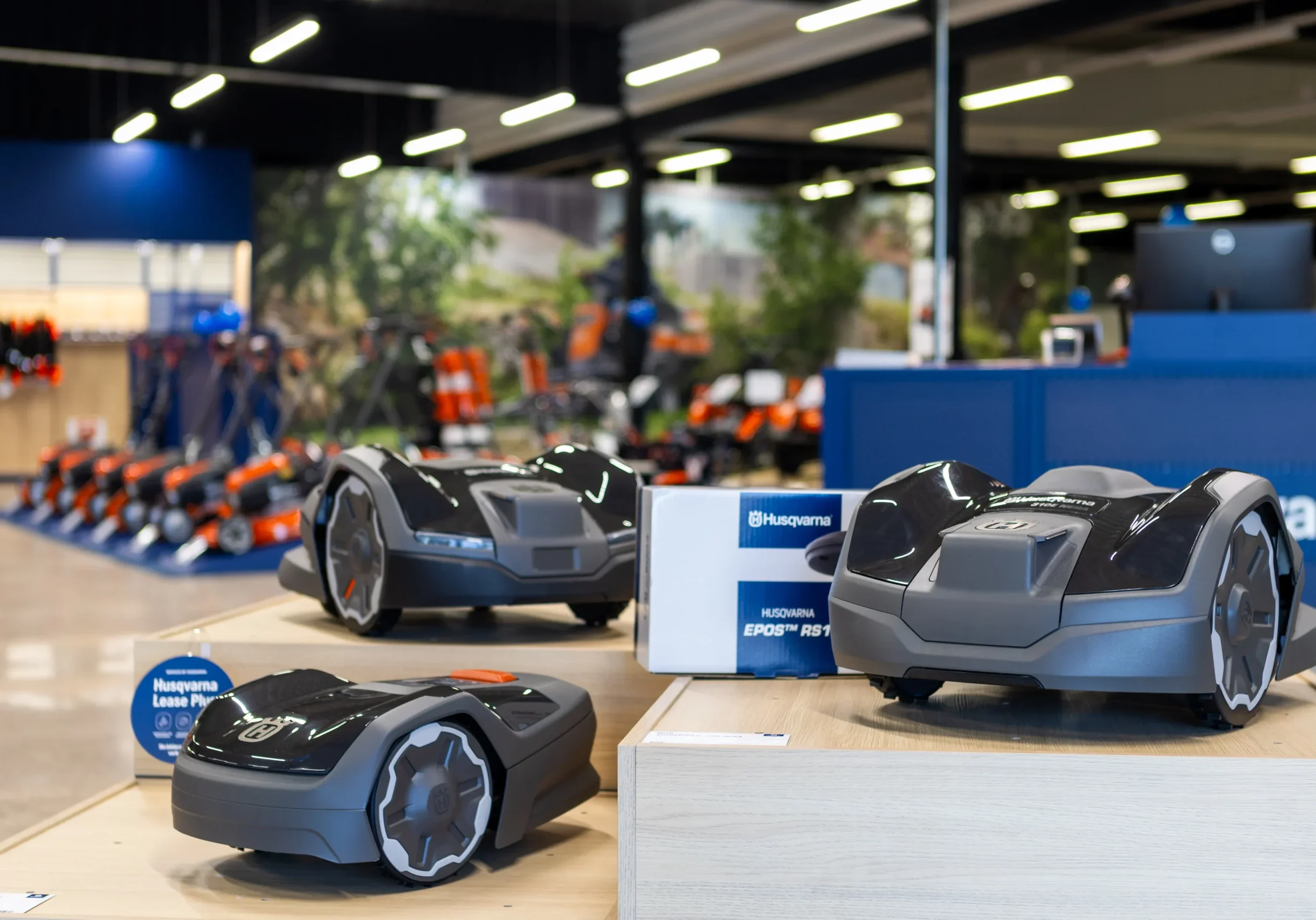 R6II1129 Tre robotgräsklippare visas på trästativ i ett väl upplyst utställningsrum, med Husqvarnas varumärke synligt och annan trädgårdsutrustning i bakgrunden.