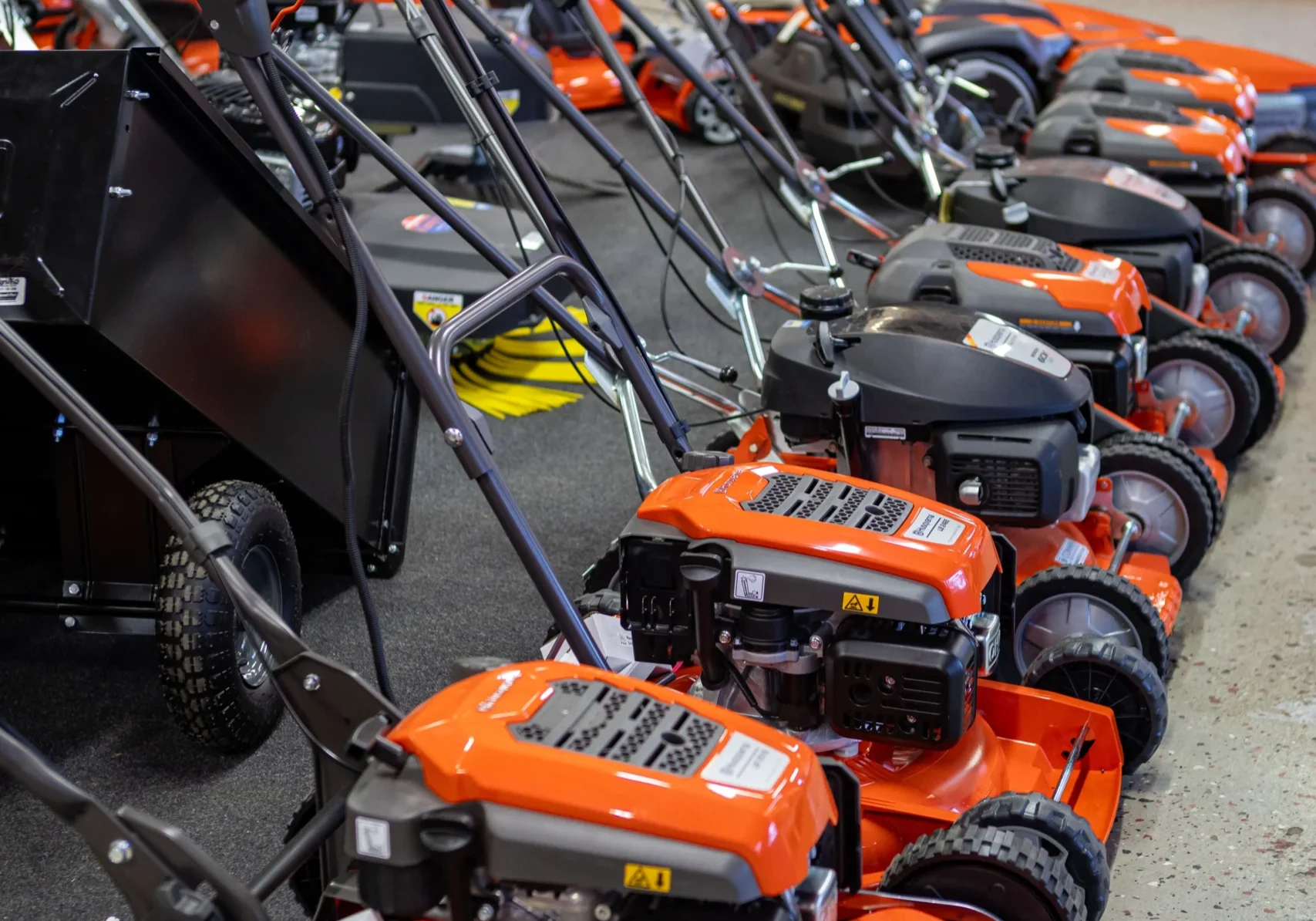 R6II6422 En rad orangefärgade gräsklippare står uppradade i en järnaffär, med olika modeller utställda på ett utställningsgolv bredvid en vägg med trädgårdsredskap - läs mer om vårt urval under "om oss.