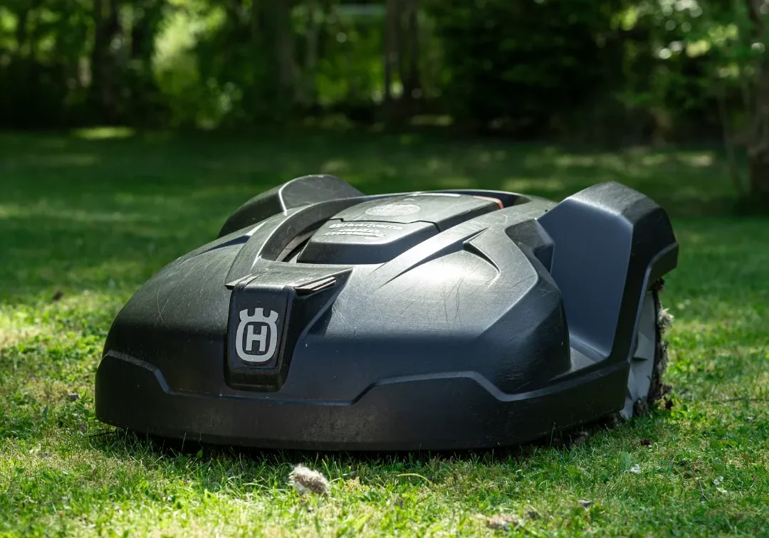 Automower A svart personlig robotgräsklippare med Husqvarna-logo står på grön gräsmatta i en solbelyst trädgård, omgiven av höga träd och frodiga växter i bakgrunden.
